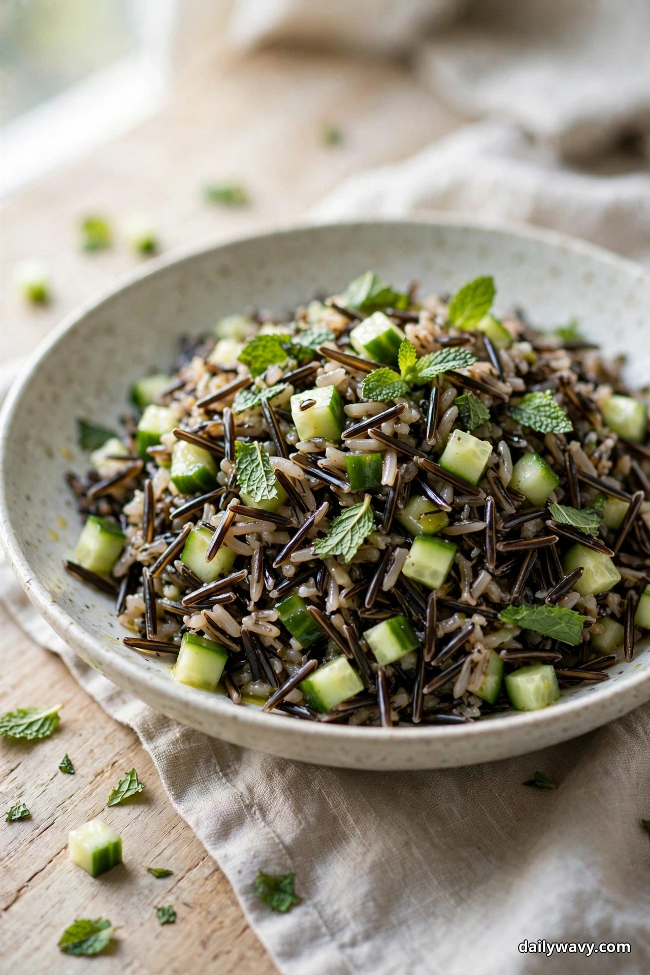 A bowl of wild rice salad with diced cucumber and fresh mint leaves sits on a linen cloth over a wooden table
